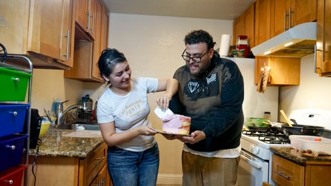Man and woman standing in a kitchen. He is holding a layer cake that she is putting pink frosting on.