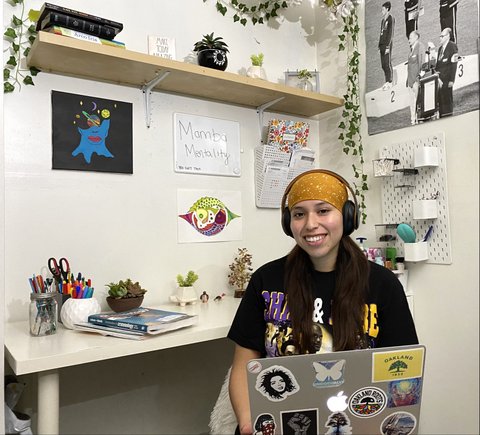 A smiling young woman wearing headphones and holding a laptop. Around her is a desk, artwork and a whiteboard with the words "Mamba Mentality."