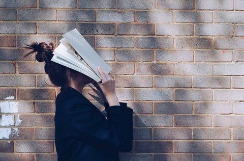 Young woman standing in front of a brick wall covering her face with a book