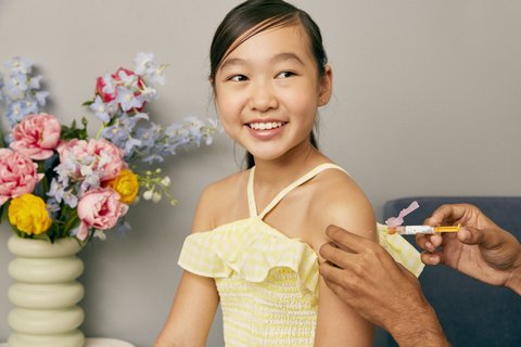 A smiling Asian girl in a yellow top getting a shot in her arm and sitting next to a bouquet of flowers.