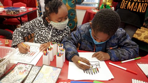 A little Black girl looking over at her brother. Both are wearing masks and coloring pictures of a kinara.