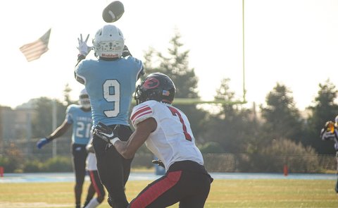 View from behind of one football player with arms outstretched ready to catch the ball as a defender moves in. The U.S. flag is visible beyond them.