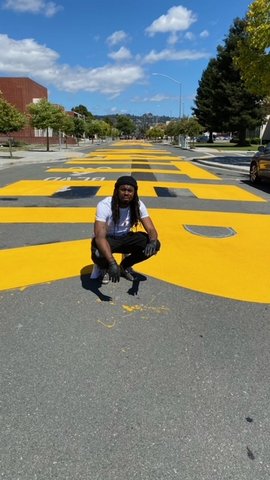 A Black man crouching in the street with large yellow letters painted behind him