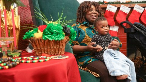 A Black woman sitting in a green chair with a baby in her lap. Around them are Christmas stockings and an arts table.
