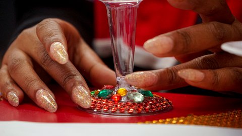 Closeup of a Black woman's hands with glittery fingernails as she glues plastic gems to the base of a small glass
