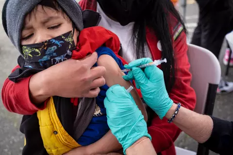 A child in a mask on someone's lap receives a vaccine shot in their arm.