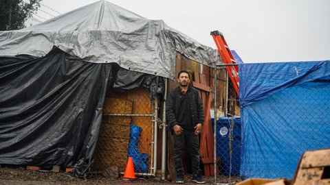 A Hispanic man stands in front of a makeshift shelter made with chain-link fencing, wood and tarps.