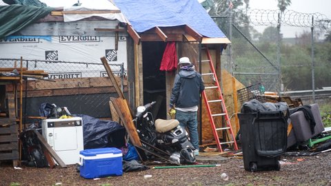 A man with his back to the camera stands near a ladder, motorcycle and other objects in front of a makeshift shelter