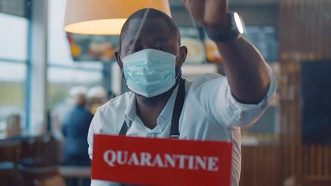Black waiter wearing safety mask hanging quarantine sign in glass door of café