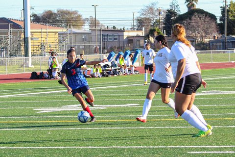 Girls' soccer player with possession of the ball and one foot raised, poised to kick, to the left of two defenders