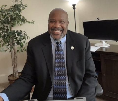 A smiling Black man in a suit with a monitor, lamp and potted plant behind him