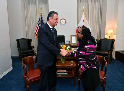 A Hispanic man and a Black woman shaking hands in a government office with U.S. and California flags