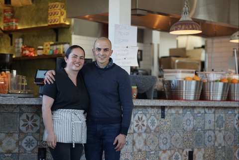 A smiling woman and man standing with his arm around her shoulder in the restaurant they own