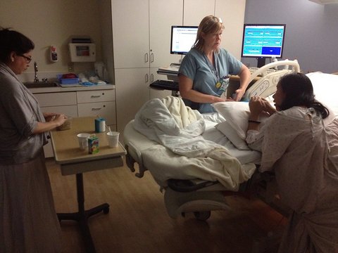 Hospital room with pregnant woman kneeling next to her bed, woman in scrubs and another woman