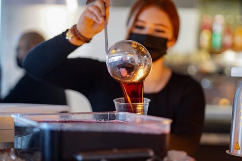 A woman in a mask pours liquid from a ladle into a cup