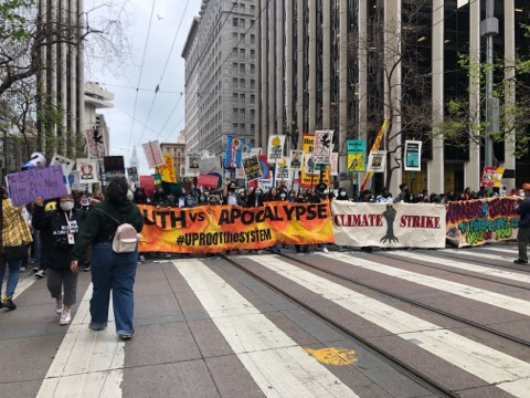 Crowd marching on downtown San Francisco street. Two banners in front read "Youth vs Apocalypse Uproot the System" and "Climate Strike"