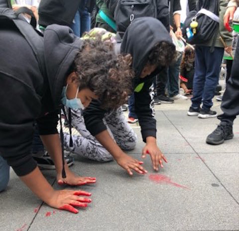 Two young men of color putting red hands on a sidewalk surrounded by a crowd
