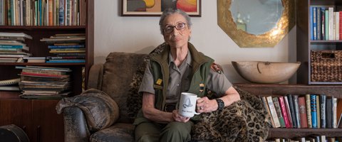 Older Black woman in park ranger uniform. She is holding a coffee cup, sitting in a cushioned chair with leopard-print blanket and surrounded by books
