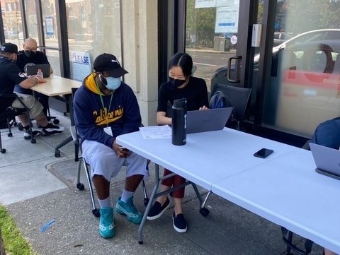 Two masked people sitting at an outdoor table in front of a laptop