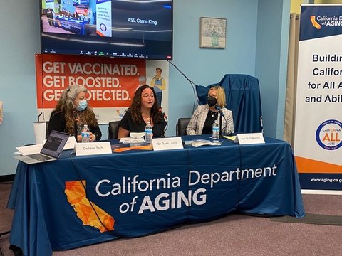 Three women at a table that says "California Department of Aging." Behind them is a sign that says "Get vaccinated, Get boosted, Get protected."
