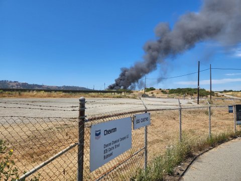 Chain-link fence with Chevron sign and barbed wire in front of large plume of smoke in the distance