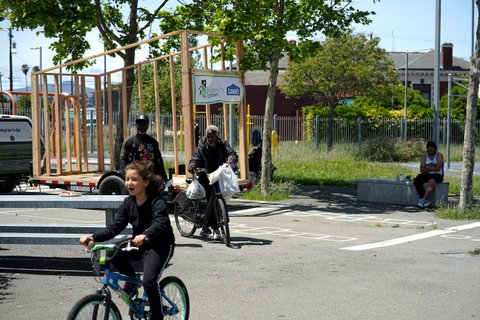 A boy riding a bike, a man walking a bike and another man walking in front of the wooden frame of a tiny home.