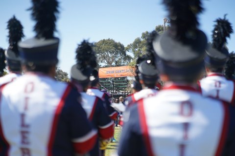 Out-of-focus marching band members seen from behind with orange banner that says "Willie Mays Junior Giants Field" clearer beyond them