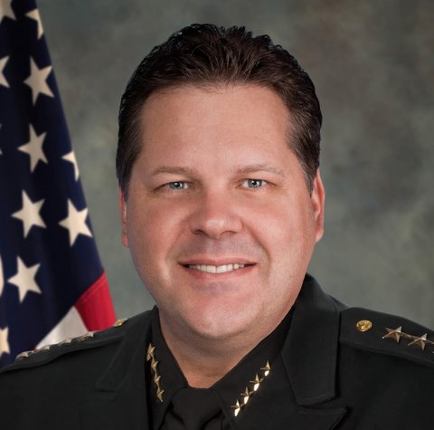 Head-and-shoulders shot of a white man in police uniform with U.S. flag over his right shoulder