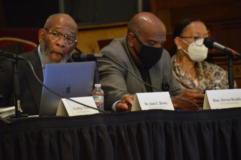 A Black man without a mask, a Black man with a mask and a Black woman with a mask sitting at a table with microphones