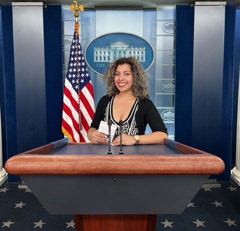 Young Latina at lectern in front of U.S. flag and White House decal