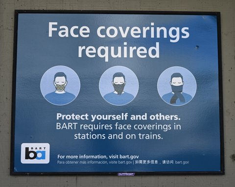 Sign with illustrations of three people with the lower halves of their faces covered that says "Face coverings required. Protect yourself and others. BART requires face coverings in stations and on trains. For more information, visit bart.gov. Para obtener más información, visite bart.gov."
