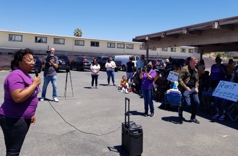 A Black woman speaks with a microphone in front of two people with video cameras. Nearby sits a crowd of people, some of which have signs protesting rent increases.