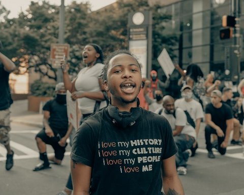 A young Black man wearing a black T-shirt that reads "i love my history. i love my culture. i love my people i love me." Out of focus behind him is a group of people, most of them on one knee.