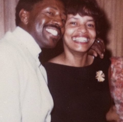 A smiling Black man and woman with his arm around her shoulders and their heads together