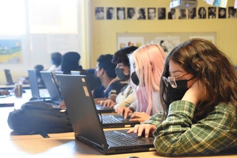 A row of high school students wearing masks and looking at laptops