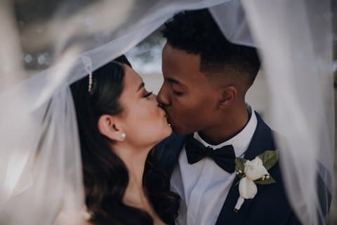 A white woman and Black man kissing. He is wearing a tuxedo. Her veil appears to be framing them