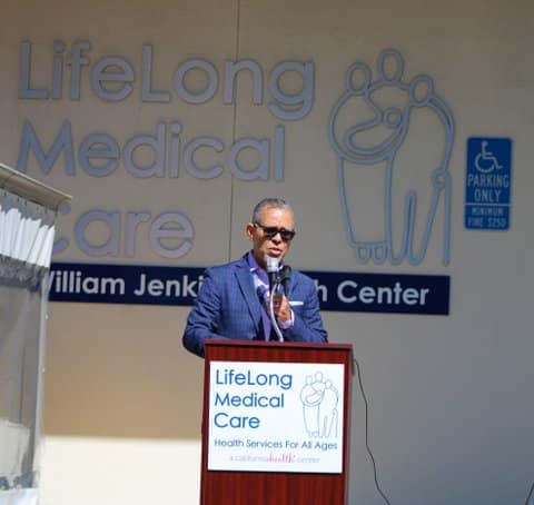 Man in sunglasses speaking at a lectern with a sign that says LifeLong Medical Care Health Services for All Ages. A California Health Center. He is standing in front of a building that says LifeLong Medical Care William Jenkins Health Center.