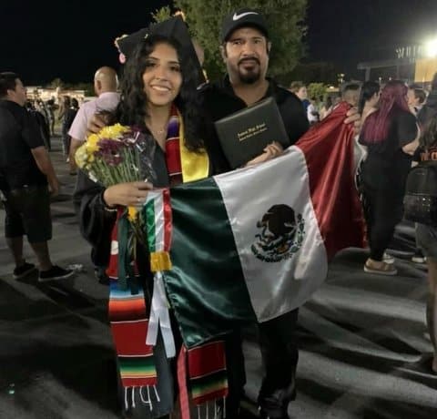 A young Latina woman dressed in cap and gown with her father. They are holding up a Mexican flag. She is also holding a bouquet of flowers and her diploma holder.