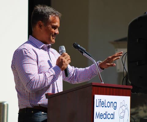 Side view of a man holding a microphone at a lectern that says LifeLong Medical