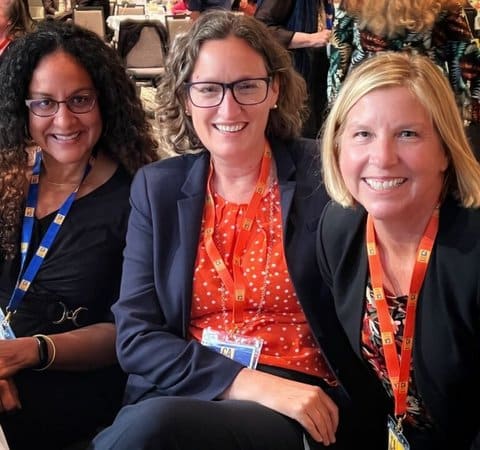 Three smiling women sitting in a row, all wearing lanyards
