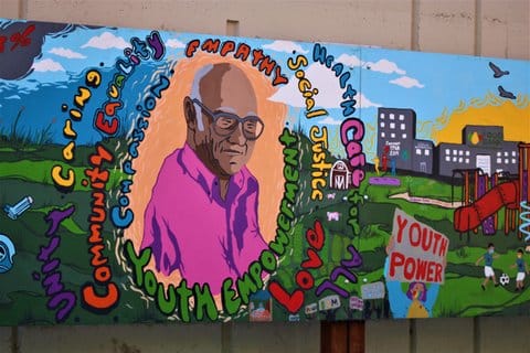 Painting of an older Black man encircled by the words empathy, social justice, youth empowerment, health care for all, unity, caring, community, equality, love. Also in the painting are a playground, two kids playing soccer, a food bank building, a farm and a person in a rainbow mask holding a youth power sign