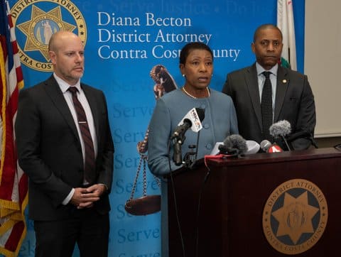 A Black woman is standing at a lectern with microphones and the Contra Costa County D.A. logo She is flanked by a white man and Black man and stands in front of a backdrop that says Diana Becton District Attorney Contra Costa County