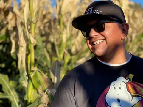 Smiling Latino man wearing black ballcap, dark sunglasses and Black Ghostbusters T-shirt. A cornfield is visible behind him.