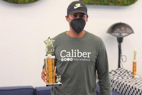 A man in a mask, baseball cap and T-shirt that says Caliber Beta Academy holding a trophy with a gold star on top