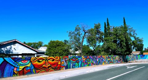 A long brick wall painted with a bright and colorful mural runs along a street and sidewalk in front of houses and trees