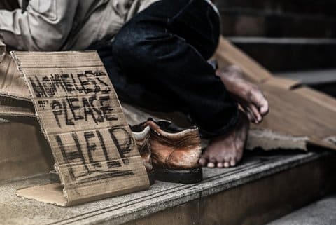 Sign that says homeless please help written on cardboard next to a pair of shoes. A Black person's bare feet and lower legs can be seen