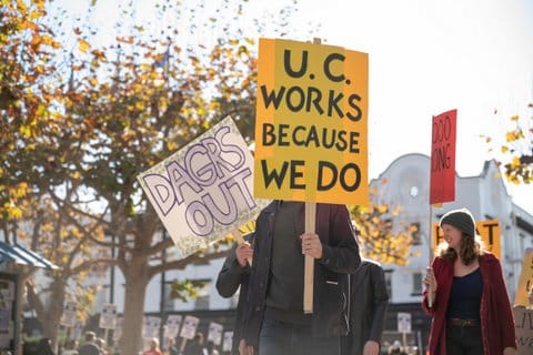 A person holds a sign in front of their face that says "U.C. works because we do" Behind them is another sign that reads "dagrs out"