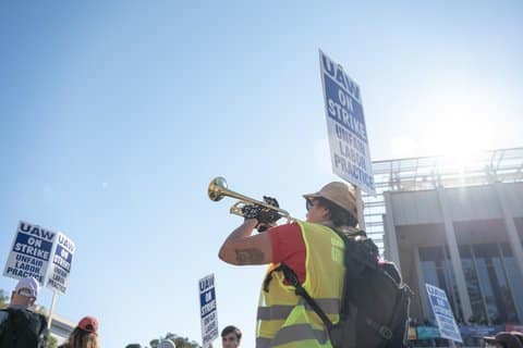 A person wearing an orange T-shirt, safety vest, helmet and backpack blows a trumpet