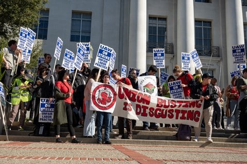 People hold a large banner that says "union busting is disgusting" among a crowd holding signs that say "UAW on strike unfair labor practices."