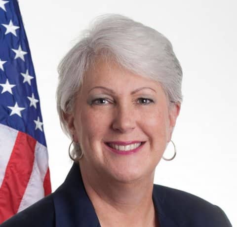 A smiling white woman with gray hair in front of a U.S. flag